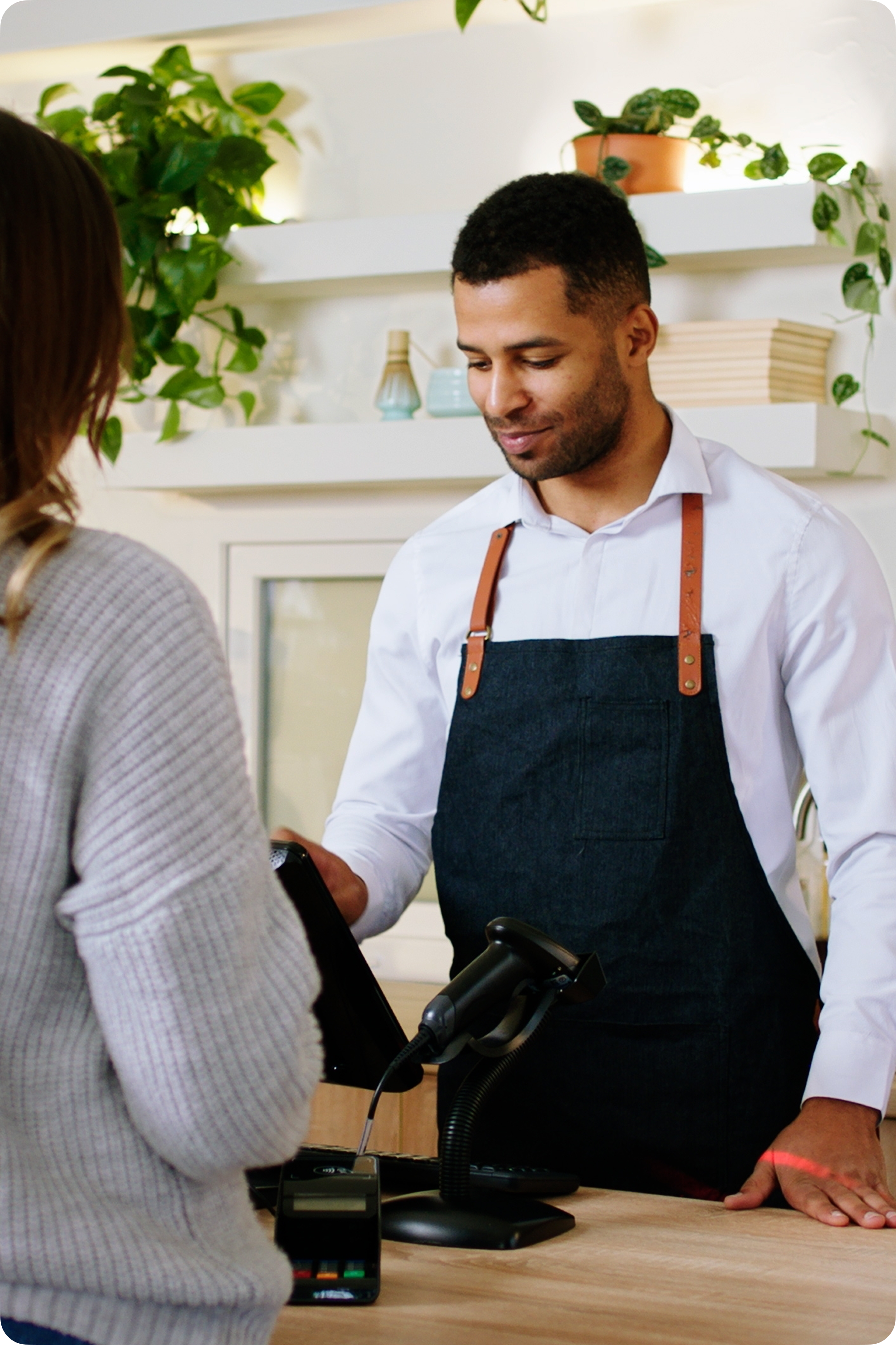 A customer stands at a checkout counter while a store employee wearing a dark apron presents a payment terminal.